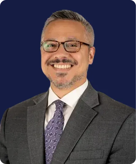 Professional middle-aged man with glasses, gray suit, and purple patterned tie, smiling against a dark blue background.