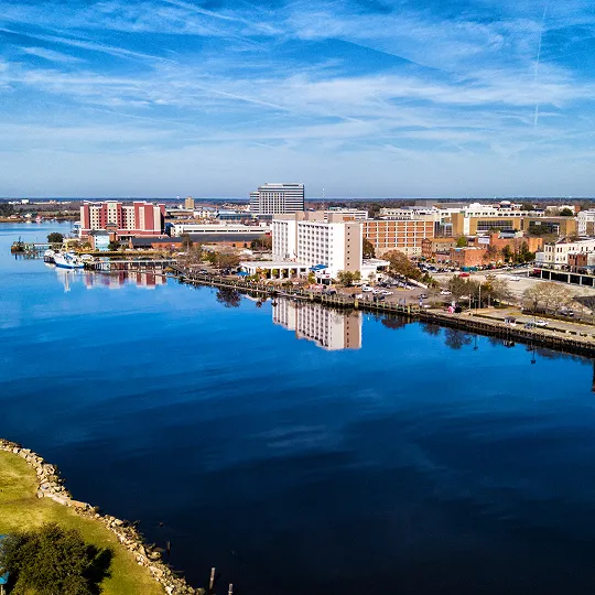 Aerial view of a waterfront cityscape with buildings reflected in calm blue water under a partly cloudy sky.
