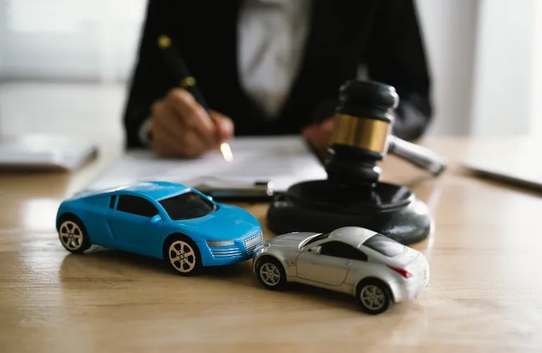 Two toy cars and a wooden gavel on a desk with a person signing documents in the background.