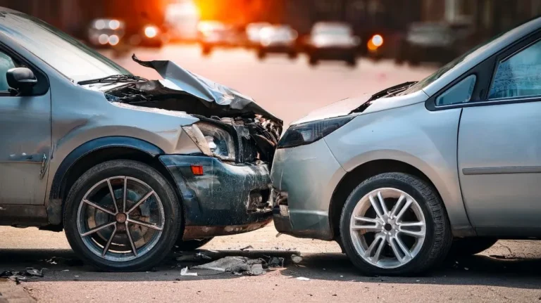 Two silver vehicles with frontal collision damage on an urban street at sunset.