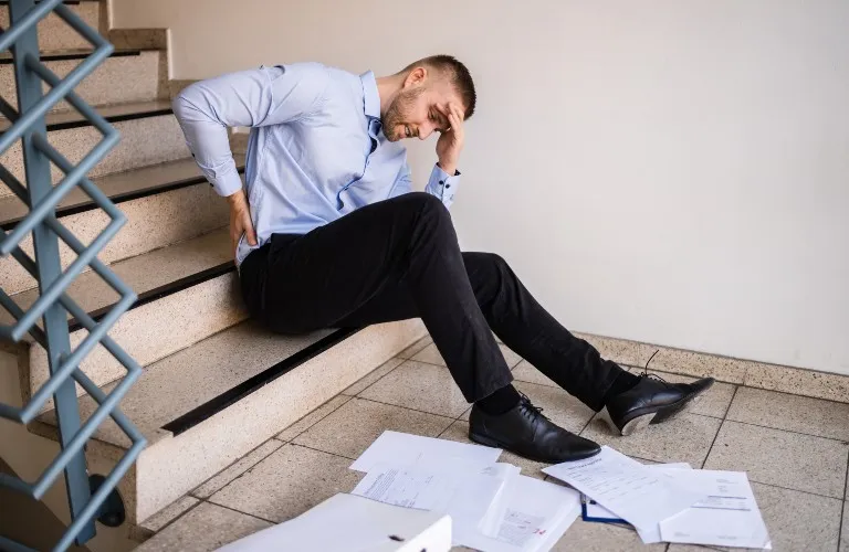 Man in blue shirt and black pants sitting on stairs, holding his back in pain, with scattered papers on the floor.