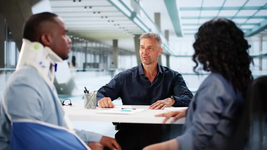 Three professionals engaged in a meeting, one wearing a neck brace and arm sling.
