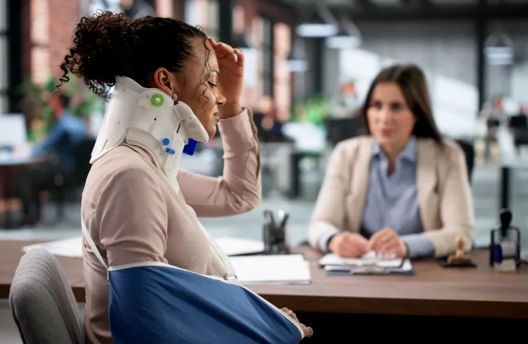 A woman wearing a neck brace and arm sling sits across a desk from a professional woman in an office.