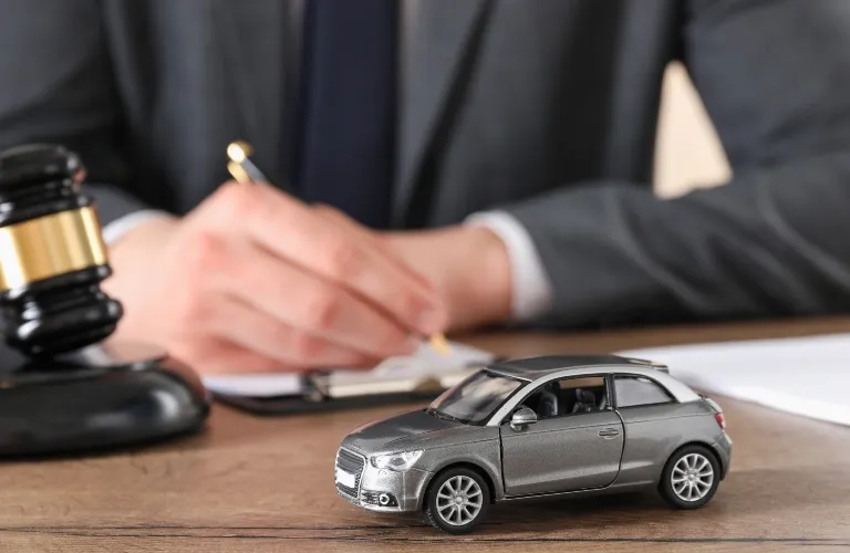A gray toy car on a wooden desk with a legal gavel and person writing in the background.
