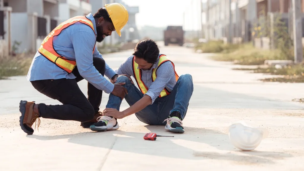 Two construction workers in safety vests, one assisting the other with a leg injury on a site.