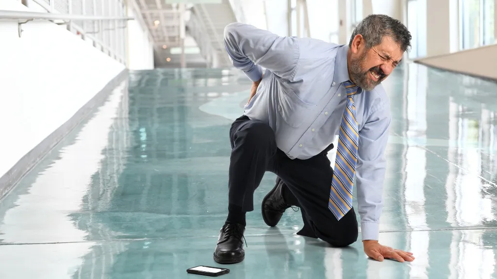 Middle-aged man in business attire kneeling on shiny floor, clutching lower back in pain.