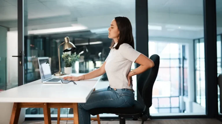 Woman in white shirt and jeans sitting at desk, holding her lower back in discomfort.