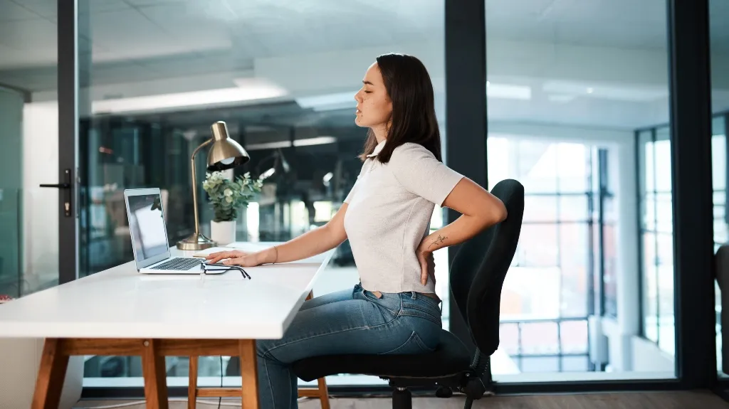 Woman in white shirt and jeans sitting at desk, holding her lower back in discomfort.