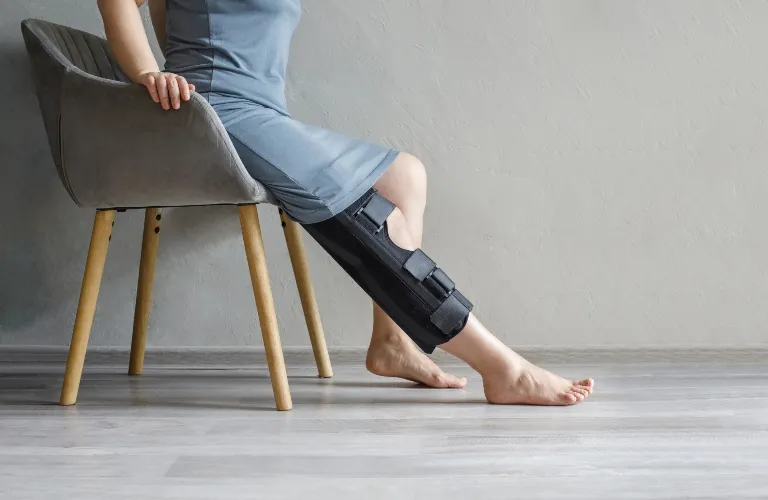 Person seated on a gray chair, wearing a leg brace on the left leg, barefoot on wooden floor.