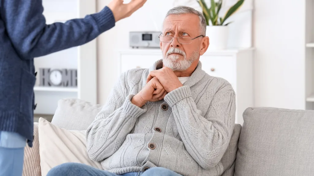 Elderly man in gray sweater sitting on couch, looking concerned during conversation.