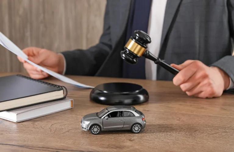 Person in suit holding gavel near toy car and documents on wooden desk.