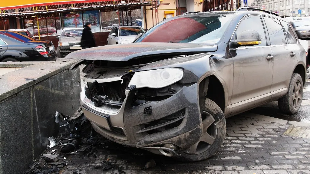 Damaged gray SUV with front-end collision, broken bumper, and raised hood parked on city street.