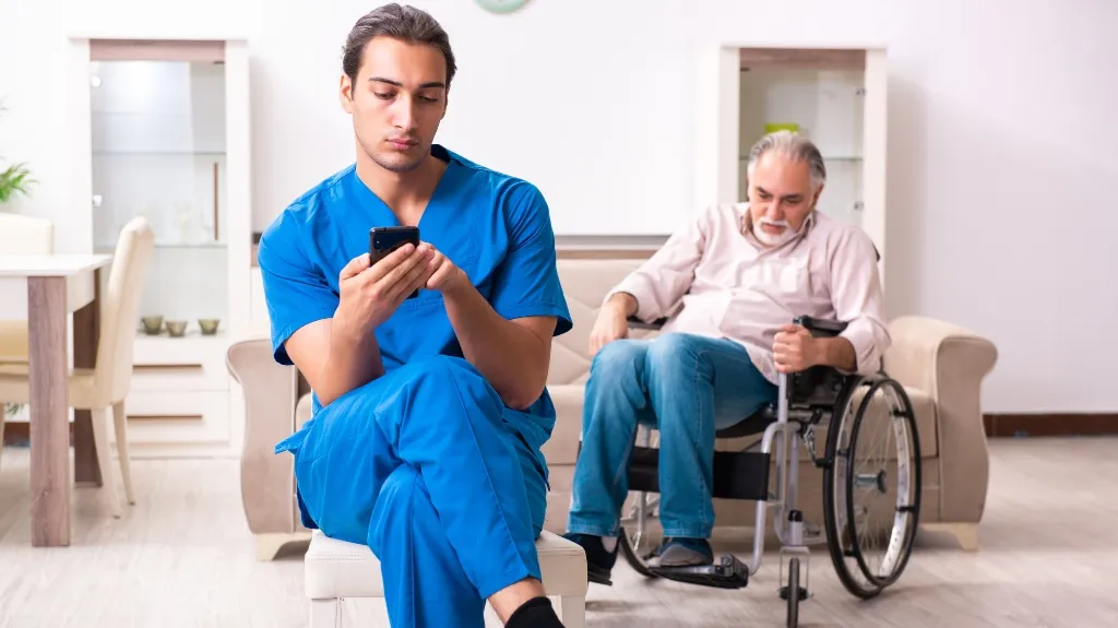Young male healthcare worker in blue scrubs using smartphone, elderly man in wheelchair in background