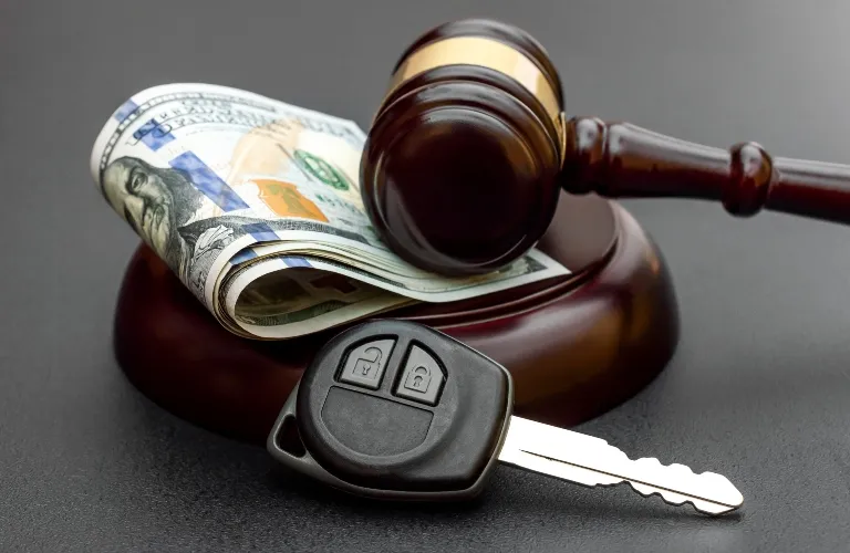 Wooden judge's gavel on block with rolled US dollar bill and black car key on black surface.