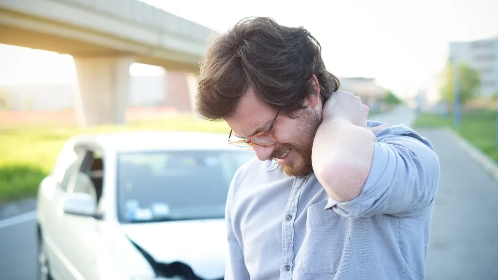 Young man with glasses holding his neck in pain near a damaged white car.