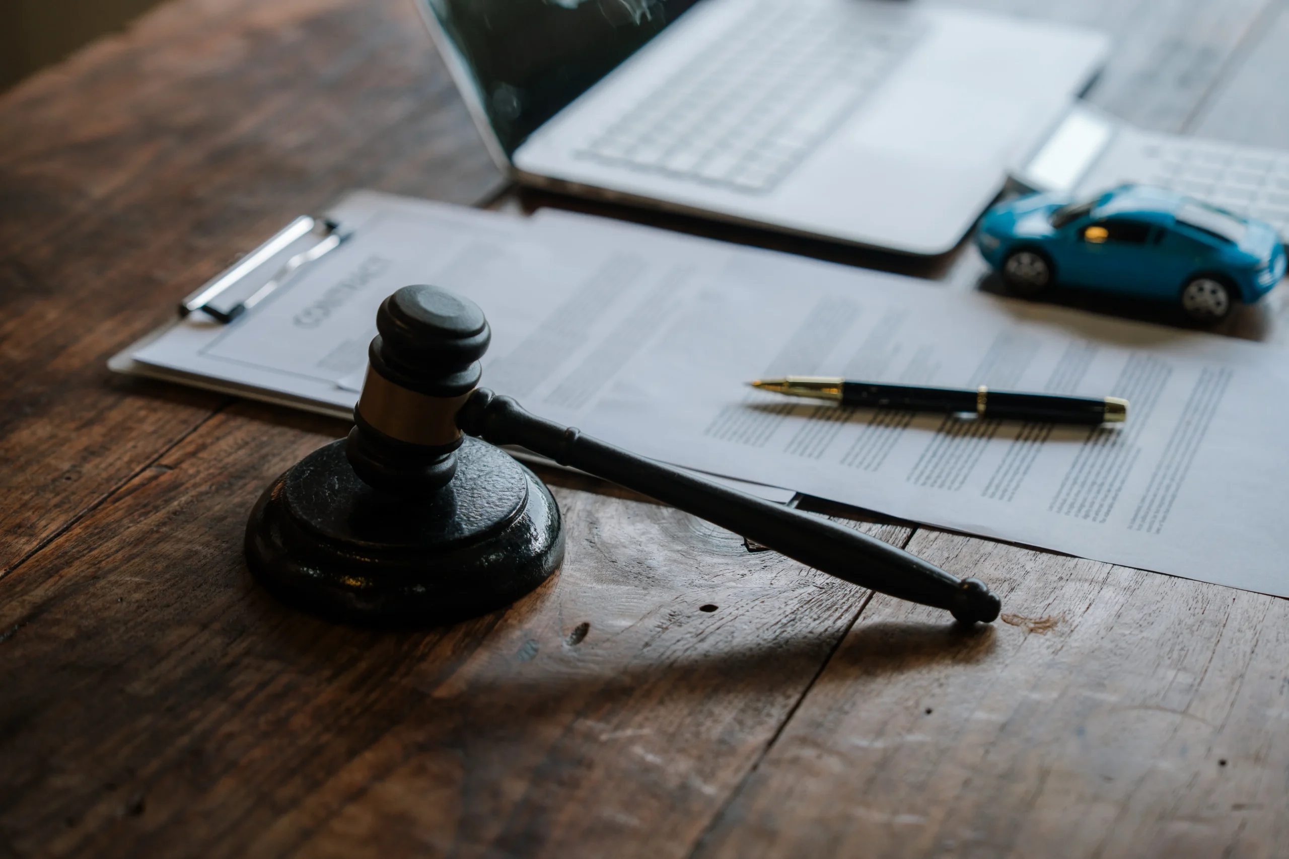 Wooden gavel, contract papers, pen, toy blue car, and laptop on rustic wooden desk.
