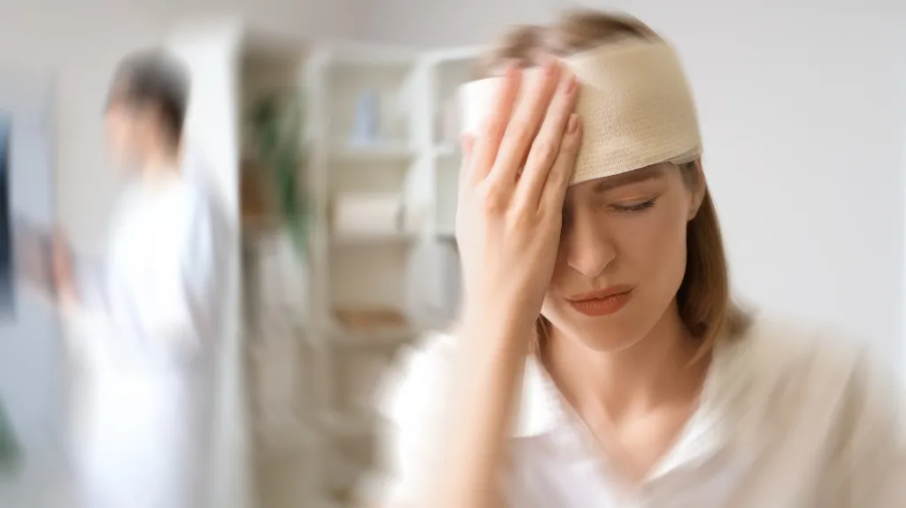 Woman with head bandage holding forehead in medical office, blurred healthcare professional in background.